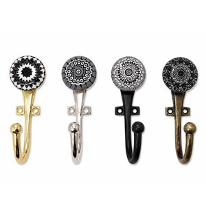 Op de afbeelding: Vier wandhaken met ronde decoratieve knoppen met zwart-witte geometrische patronen. Elke haak heeft een eigen metalen afwerking: gepolijst goud, glanzend zilver, matzwart en een verouderde bronskleur, met een kogelvormig uiteinde.