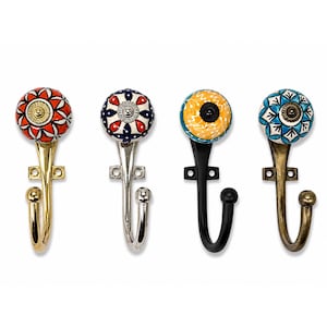 Könnte beinhalten: Vier Wandhaken mit runden Keramikknöpfen, die farbenfrohe Blumen- und geometrische Muster aufweisen. Jeder Haken hat ein eigenes Metallfinish, darunter poliertes Gold, Silber, Mattschwarz und Antikbronze, mit einem geschwungenen Arm.