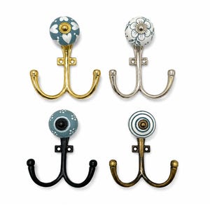 Puede incluir: Cuatro ganchos de pared dobles con pomos de cerámica decorativos. El gancho superior izquierdo es dorado con un patrón de corazones azul y blanco. El superior derecho es plateado con un diseño floral. El inferior izquierdo es negro con un pomo azul de puntos y el inferior derecho es bronce con un pomo de rayas azul y blanco.