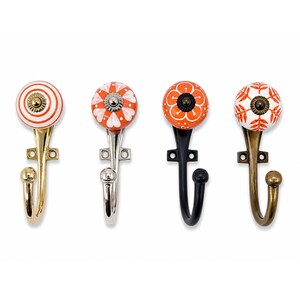 Op de afbeelding: Vier wandhaken met ronde keramische knoppen in oranje en witte patronen. Elke haak heeft een eigen metalen afwerking, waaronder gepolijst goud, zilver, matzwart en antiek messing, met een montageplaat en een enkele gebogen haak.