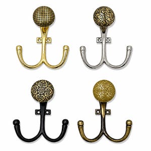 May include: Four double-pronged wall hooks featuring decorative round knobs. The top left hook is gold with a woven pattern, the top right is silver with an intricate floral design, the bottom left is black with a floral pattern, and the bottom right is antique brass with a floral motif.