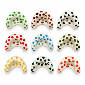 May include: Nine crescent-shaped ceramic objects arranged in a grid, each featuring a white base with hand-painted polka dots in various colours including green, brown, black, red, blue, olive, yellow, dark green, and orange.