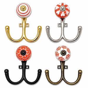 May include: Four double-pronged wall hooks with decorative ceramic knobs. The top left hook is gold with a red and white striped knob. The top right is silver with a red and white heart pattern. The bottom left is black with a red floral knob. The bottom right is bronze with a white and orange leaf pattern.