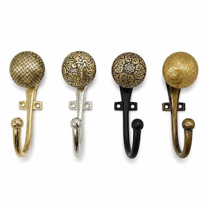 Puede incluir: Cuatro ganchos de pared con pomos esféricos, cada uno con un patrón texturizado distinto. Los ganchos tienen acabados en oro metálico, plata, negro y bronce antiguo, con una placa de montaje y un brazo curvo con un remate esférico.