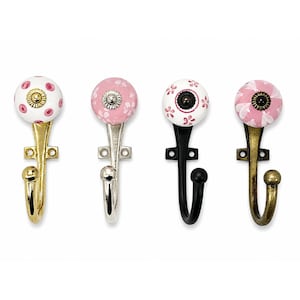 Pode incluir: Quatro ganchos de parede com puxadores de cerâmica redondos. O primeiro tem uma base branca com pontos cor-de-rosa e acabamento dourado. O segundo é cor-de-rosa com padrões florais brancos e acabamento prateado. O terceiro é branco com flores cor-de-rosa e acabamento preto. O quarto é cor-de-rosa com pétalas brancas e acabamento em latão antigo.