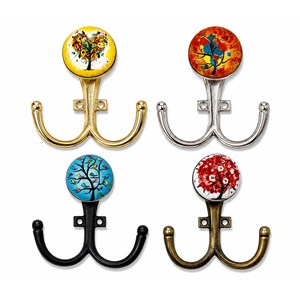 May include: Four double-pronged wall hooks featuring circular decorative knobs. The top left hook is gold with a yellow tree design, the top right is silver with a red tree, the bottom left is black with a blue tree, and the bottom right is bronze with a red floral tree.