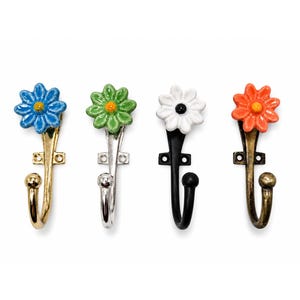 Puede incluir: Cuatro ganchos de pared con cabezas de flores de cerámica en azul, verde, blanco y naranja. Cada gancho tiene una base de metal con placa de montaje y un brazo curvo que termina en una punta de bola. Los acabados metálicos incluyen oro, plata, negro y bronce.