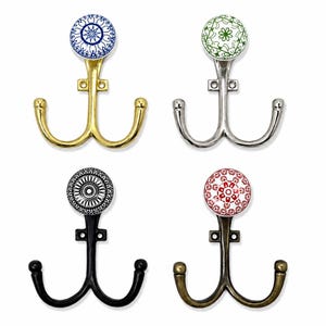 Puede incluir: Cuatro ganchos de pared dobles con pomos de cerámica decorativos. Los ganchos tienen acabados metálicos en oro, plata, negro y bronce. Cada pomo muestra un patrón circular único en azul, verde, negro o rojo sobre un fondo blanco.