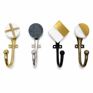 May include: Four wall hooks with distinct decorative heads. The first is a white marble circle with a gold cross. The second is a dark gray textured circle on a silver hook. The third is a square with white and gold halves on a black hook. The fourth is a white and gold circle on a brass hook.