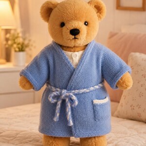 May include: A plush tan teddy bear wearing a light blue knitted robe with a white braided tie and a white pocket trim. The bear has dark eyes, a black nose, and a white shirt collar visible underneath the robe. The fabric has a soft texture.