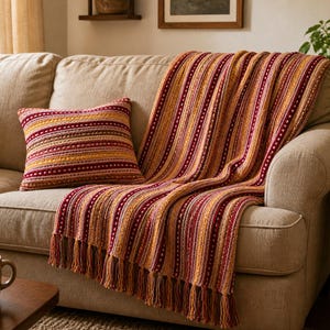 Könnte beinhalten: Eine gestrickte Wohndecke und ein passendes quadratisches Kissen mit vertikalem Streifenmuster in den Farben Burgunderrot, Senfgelb und Creme. Die Decke hat ein strukturiertes Strickmuster und ist an der Kante mit langen Fransen versehen.