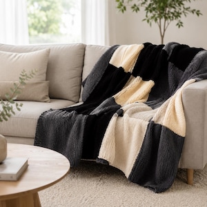 Könnte beinhalten: Eine grob gestrickte Wohndecke mit einem großflächigen Schachbrettmuster aus abwechselnden Blöcken in Creme, Anthrazit und Schwarz. Die Decke besteht aus dickem, strukturiertem Garn und liegt über der Armlehne und der Sitzfläche eines Sofas.