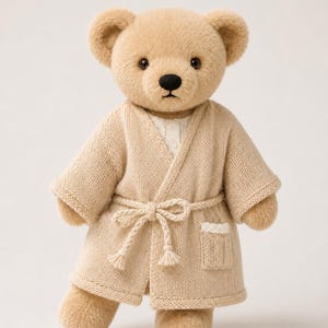 Può includere: Un orsacchiotto di peluche con pelliccia marrone chiaro, occhi scuri e naso nero. L'orso indossa un accappatoio in maglia beige con cintura abbinata e una piccola tasca con bordo bianco, sopra una camicia bianca dalla trama visibile.
