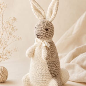 May include: A two-toned knitted rabbit toy featuring a cream-colored front and a light brown back. The rabbit has long upright ears, dark embroidered eyes and nose, and a small white satin ribbon tied around its neck. A fluffy white tail is visible.