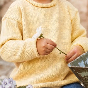 May include: A pale yellow long-sleeved knit sweater featuring a rolled collar and ribbed cuffs. The sweater has a simple, plain texture and is worn with blue denim jeans. The garment is constructed with a classic pullover design and a straight hem.