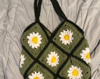 May include: Handmade crochet tote bag featuring a daisy pattern. The bag is made of green yarn with white and yellow daisy appliqués. The bag has a dark green border and matching handles. The bag is designed to be carried over the shoulder.