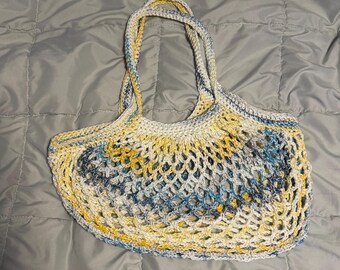 May include: A crocheted tote bag in shades of yellow, blue, and white. The bag features a net-like design with two handles. The bag is sitting on a gray surface.