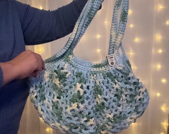 May include: A handmade crochet bag in shades of blue, green, and white. The bag features a unique open weave design and long straps for carrying. The bag is held up by a person.