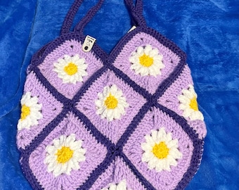May include: A handmade crochet tote bag in a daisy pattern. The bag features a lilac background with white and yellow daisy appliqués. The bag has a dark purple border and handles, and is set against a blue background.