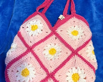 May include: A handmade crochet tote bag featuring a daisy pattern. The bag is primarily pink with white and yellow daisy accents. The straps and trim are a vibrant fuchsia color. The bag is designed with a grid pattern.