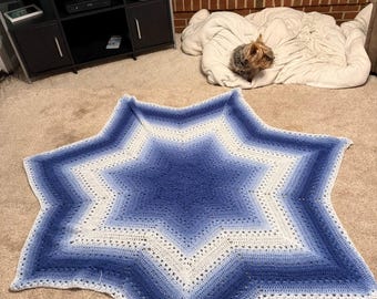 May include: A large, star-shaped crochet blanket in shades of blue and white. The blanket features a gradient design, transitioning from a deep blue center to lighter blue and white edges. The blanket is handmade and appears soft.