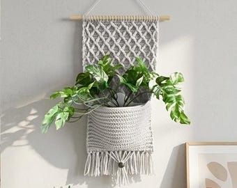 May include: A grey macramé wall planter with a woven diamond pattern and a fringed bottom. The planter holds a green leafy plant. The planter is suspended from a wooden dowel and a white cord.