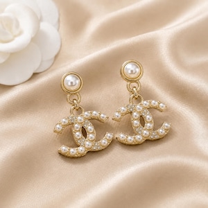 May include: A pair of gold-tone drop earrings featuring a dangling interlocking CC logo design. Each earring is embellished with small white faux pearls and clear crystals, topped with a single larger round faux pearl stud post.