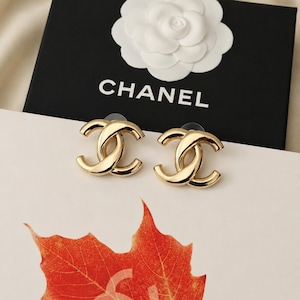 May include: A pair of gold-toned earrings featuring the interlocking CC logo. The earrings are positioned in front of a black box with the white text CHANEL and a white camellia flower emblem, resting above a vibrant orange autumn leaf.