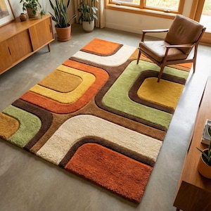 Op de afbeelding: Een rechthoekig vloerkleed met een herhalend patroon van afgeronde geometrische vormen in tinten oranje, geel, olijfgroen, bruin en crème. Het kleed heeft een dikke, getextureerde pool en ligt op een gladde, lichtgrijze betonnen vloer.