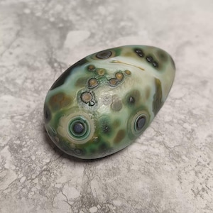 May include: A smooth, oval-shaped stone featuring a polished surface with a mottled pattern of sage green, olive, and dark forest green tones. The stone displays distinct circular eye-like markings and small brown spots scattered across its surface.