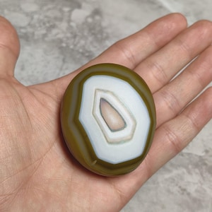 May include: A polished, rounded agate stone featuring concentric bands of olive green and white, surrounding a central, smooth, light-coloured core. The stone displays a distinct, layered pattern with a smooth, glossy surface texture throughout.