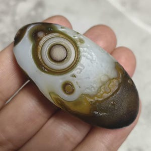 May include: An oval-shaped agate stone featuring a prominent, centred eye-like pattern with concentric white and brown rings. The surface displays a smooth, polished texture with a colour palette of milky white, deep brown, and olive green.