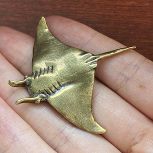 May include: A bronze-colored manta ray pendant. The pendant features a detailed design of the ray's body, wings, and tail. The wings are spread wide, and the tail extends to the right. The pendant is likely made of metal and has a vintage or antique appearance.