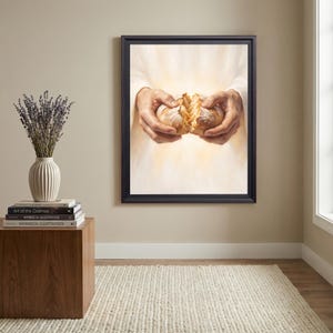 May include: A framed wall art print depicting two hands breaking a loaf of bread against a bright, glowing white background. The artwork is displayed in a black frame and features a soft, radiant light emanating from behind the bread.
