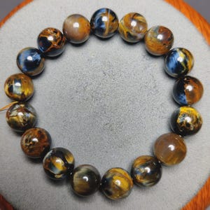 May include: A circular bracelet composed of smooth, polished spherical beads. The beads feature a chatoyant pattern with swirling bands of deep blue, golden brown, and black, creating a reflective, multi-colored appearance throughout the piece.