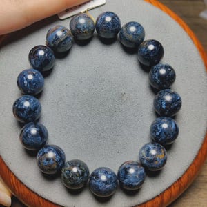 May include: A circular bracelet composed of polished, spherical beads made of blue pietersite stone. Each bead displays a unique, swirling chatoyant pattern with deep navy, royal blue, and subtle golden-brown inclusions throughout the polished surface.