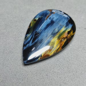 May include: A teardrop-shaped pietersite gemstone cabochon featuring a polished surface with swirling patterns of deep blue, black, and golden-yellow chatoyant fibers. A small white sticker with the black printed text 40/54E is attached.