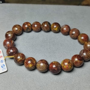 May include: A bracelet composed of polished, spherical beads in shades of deep brown, reddish-brown, and golden yellow. The beads are strung together in a circular arrangement, and a small white rectangular tag with blue and red text is attached.