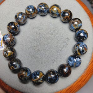 Puede incluir: Una pulsera circular compuesta por cuentas esféricas pulidas que presentan un patrón de remolinos en colores azul profundo, negro y marrón dorado. Las cuentas están ensartadas en un bucle continuo, mostrando un acabado brillante.
