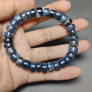 Puede incluir: Una pulsera compuesta por numerosas cuentas cilíndricas con un patrón jaspeado pulido en azul profundo y gris oscuro. Las cuentas están ensartadas en una formación circular, mostrando una superficie lisa y reflectante con texturas de piedra natural.