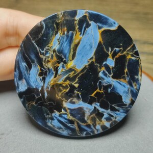 May include: A circular polished gemstone cabochon featuring a complex pattern of deep black, vibrant blue, and golden-brown streaks. The stone displays a chatoyant, fibrous texture with high-contrast colour variations across its smooth surface.