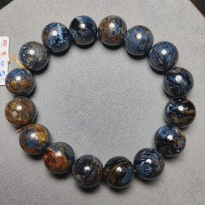 Op de afbeelding: Een cirkelvormige armband bestaande uit vijftien gepolijste, bolvormige kralen. De kralen hebben een wervelend, gemarmerd patroon van diep marineblauw, leigrijs en aardse bruintinten met een glanzende, reflecterende afwerking.