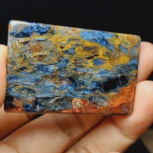 May include: A rectangular polished gemstone cabochon featuring a swirling pattern of deep blue, golden yellow, and reddish-brown mineral inclusions. The surface is smooth and glossy, displaying a complex, fibrous texture throughout the stone.
