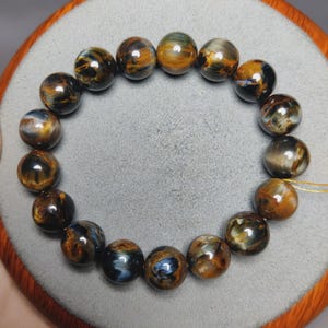 May include: A circular bracelet composed of polished, spherical beads made of tiger's eye stone. The beads feature a chatoyant pattern with swirling bands of deep golden-brown, dark charcoal, and iridescent blue tones, strung together on a thin cord.