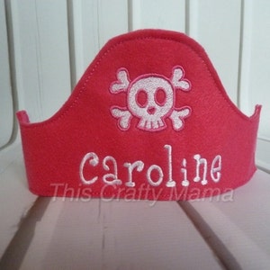 May include: A pink felt pirate hat with a white skull and crossbones and the name "Caroline" embroidered on it.
