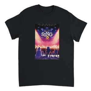May include: Black t-shirt featuring a colorful movie poster for "Sing 2." The poster shows characters in silhouette against a stage with bright lights and the movie title. The shirt is made of a soft material.