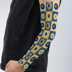 May include: An arm sleeve featuring a repeating checkered pattern of yellow and blue squares. The yellow squares contain dark blue sun and moon icons, while the blue squares feature yellow sun and moon icons, creating a celestial design.