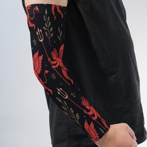 May include: A black arm sleeve featuring a repeating pattern of red winged devil figures holding tridents, interspersed with thin gold-colored botanical sprigs. The fabric is form-fitting and covers the forearm and elbow area completely.