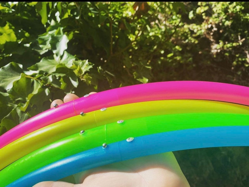 May include: Four colourful plastic hula hoops stacked on top of each other. The hoops are pink, yellow, green, and blue. The hoops are decorated with silver metal accents.
