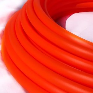 May include: A close-up of a stack of bright orange hula hoops. The hoops are made of a flexible plastic material and are stacked on top of each other.